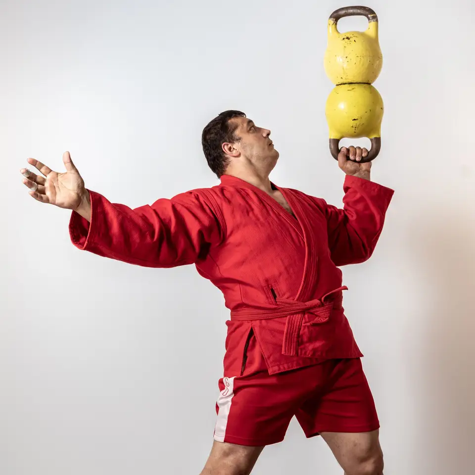 Male athlete in red uniform showcasing strength with double kettlebell lift on white background.