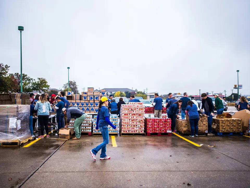 Volunteers handing out boxed food from pallets in a large parking lot on an overcast day