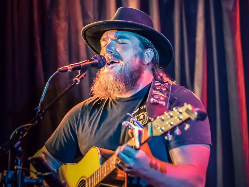 Rowan Hale playing an acoustic guitar in front of red curtains