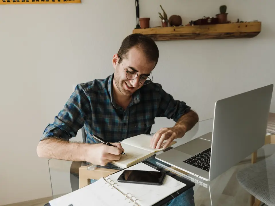 Man taking notes beside a laptop and phone at a home desk