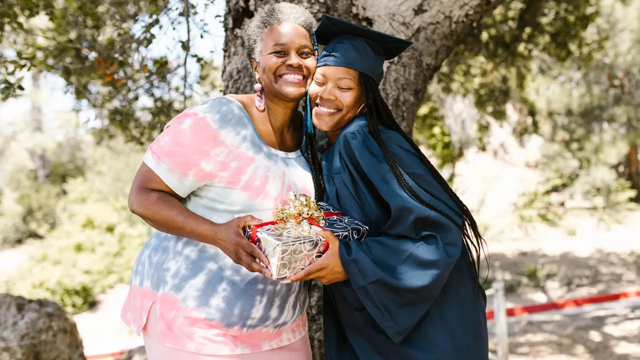 Graduate hugging her mother, celebrating with a gift under a tree