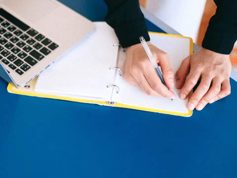 Hands writing notes in a notebook next to a laptop