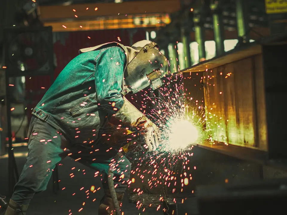 A focused welder wearing protective gear as bright sparks fly in a factory setting.